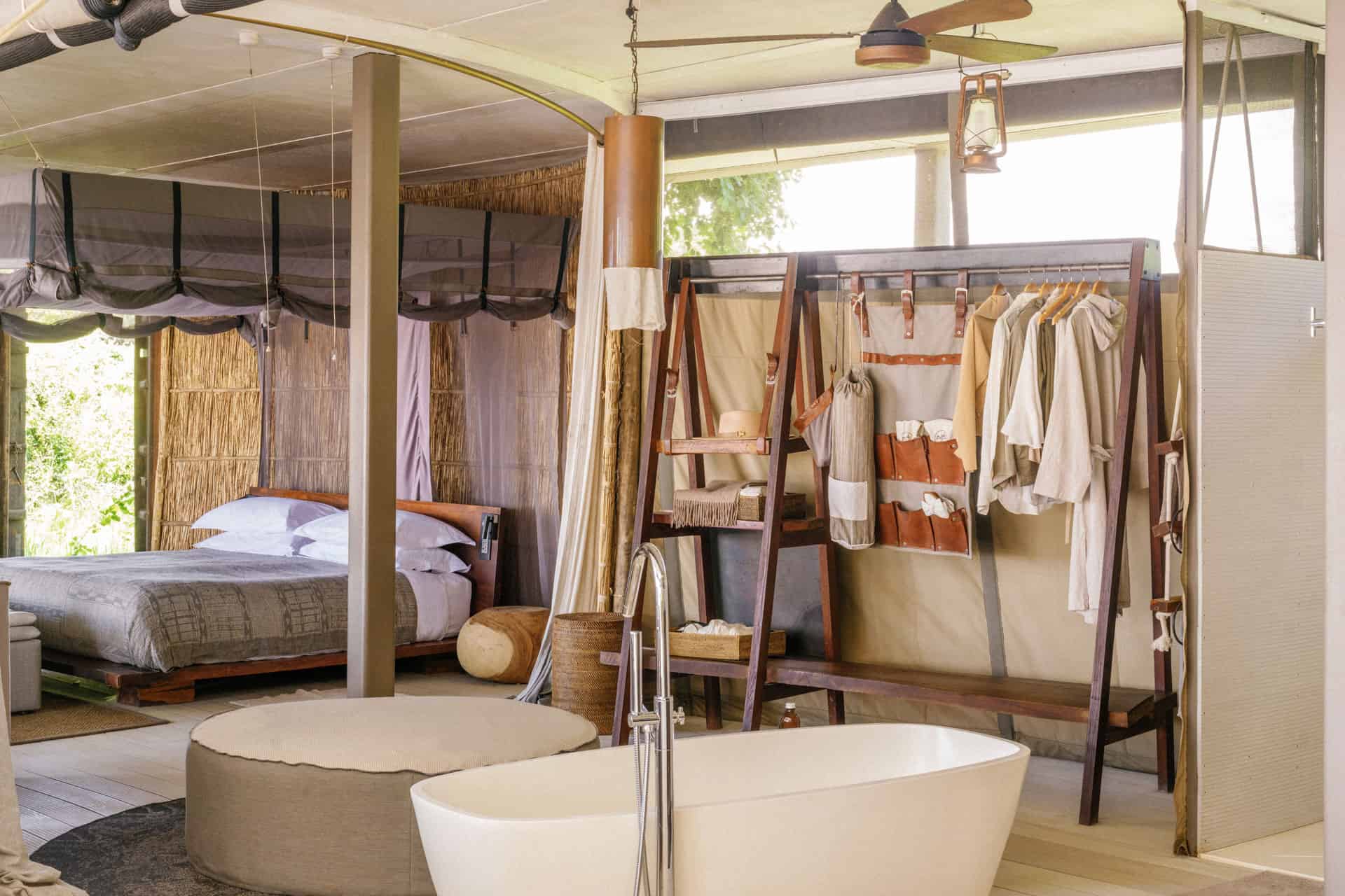 A spacious room is visible featuring a neatly made bed, a modern freestanding bathtub, and a wooden clothing rack with garments, all designed with natural materials and abundant light.