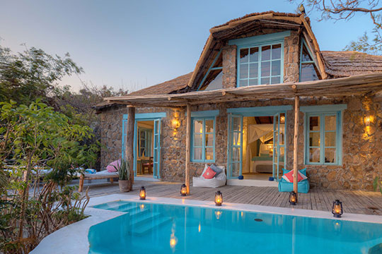 A rustic stone house featuring a thatched roof and light blue trim stands beside a swimming pool and wooden deck illuminated by lanterns, surrounded by lush greenery under a clear sky.
