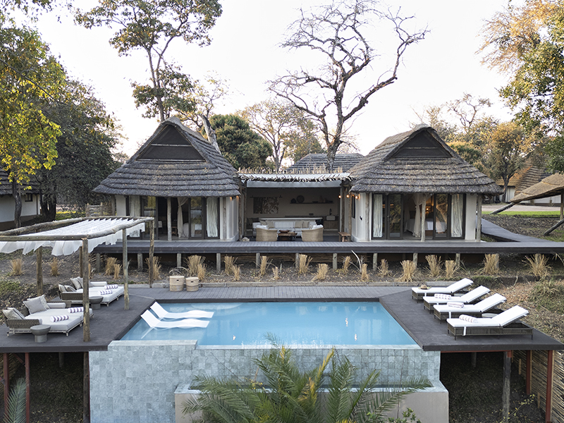 Two thatched roof bungalows with an open air central lounge stand on an elevated wooden deck featuring a swimming pool, several lounge chairs, and shaded seating, all surrounded by trees.